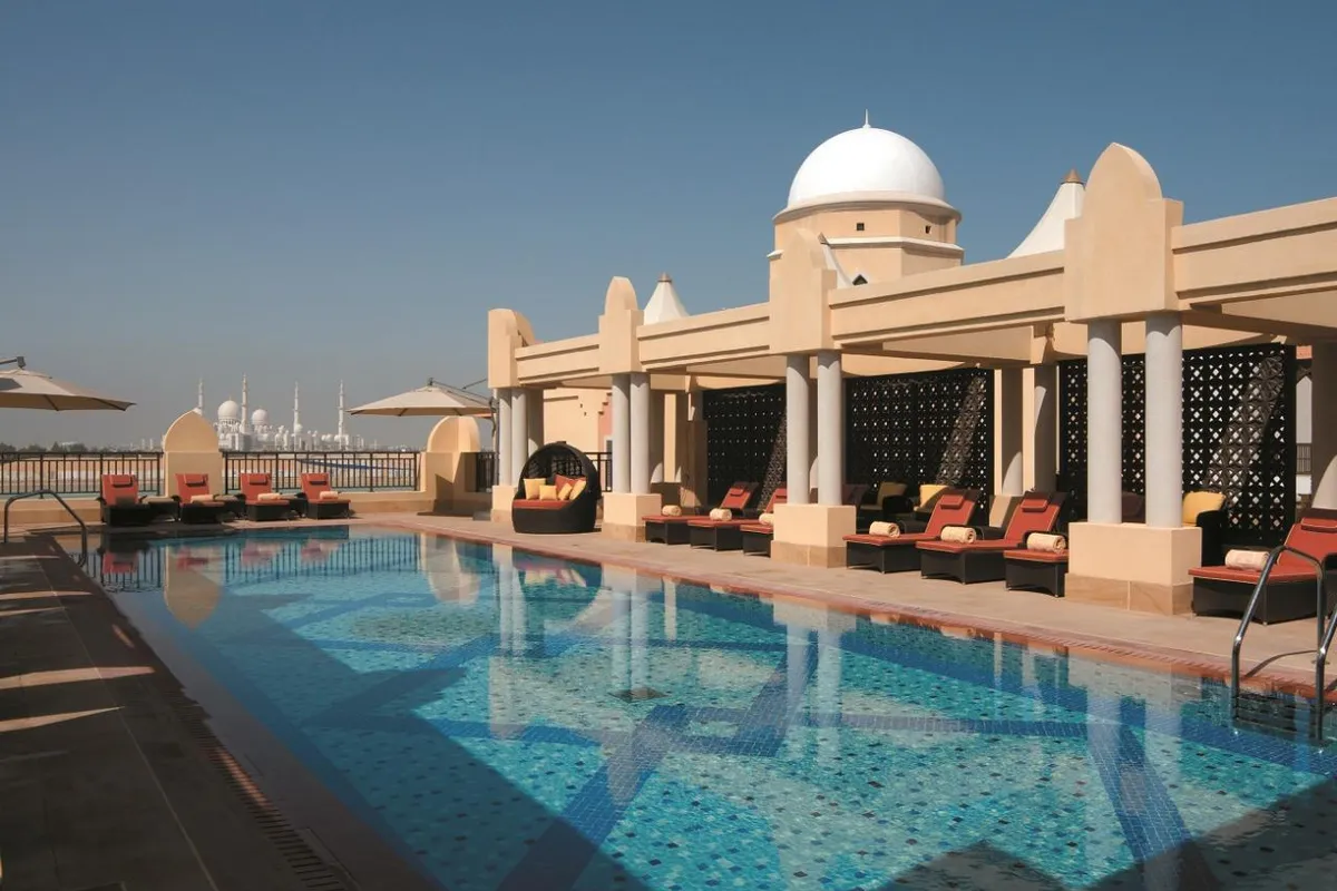Basen na tarasie hotelu Shangri-La Qaryat Al Beri w Abu Zabi, leżaki pod arkadami z kopułą, a w oddali białe kopuły i minarety Wielkiego Meczetu