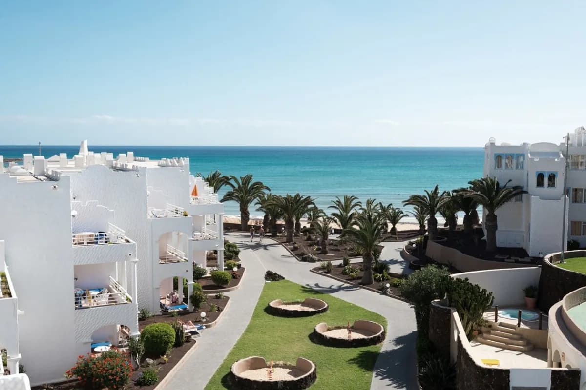 Białe budynki hotelu i aleja z palmami prowadząca do plaży w Costa Calma na Fuerteventurze, w tle turkusowy ocean