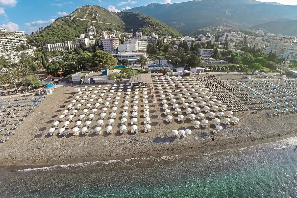 Szeroka plaża w Bečići koło Budvy z rzędami białych parasoli, za nią hotele i zalesione góry schodzące ku morzu