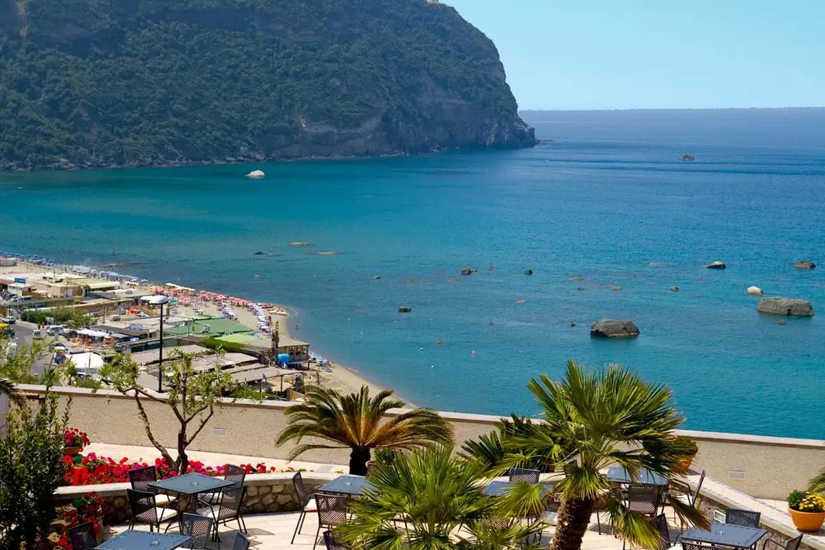 Widok z tarasu hotelu Royal Palm w Forio na Ischii: piaszczysta plaża z parasolami, turkusowa zatoka ze skałami i zalesiony cypel