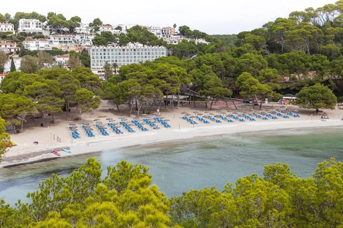 Plaża w zatoce Cala Galdana na Minorce: jasny piasek z niebieskimi leżakami, sosny schodzące do spokojnej wody i hotele na zboczu
