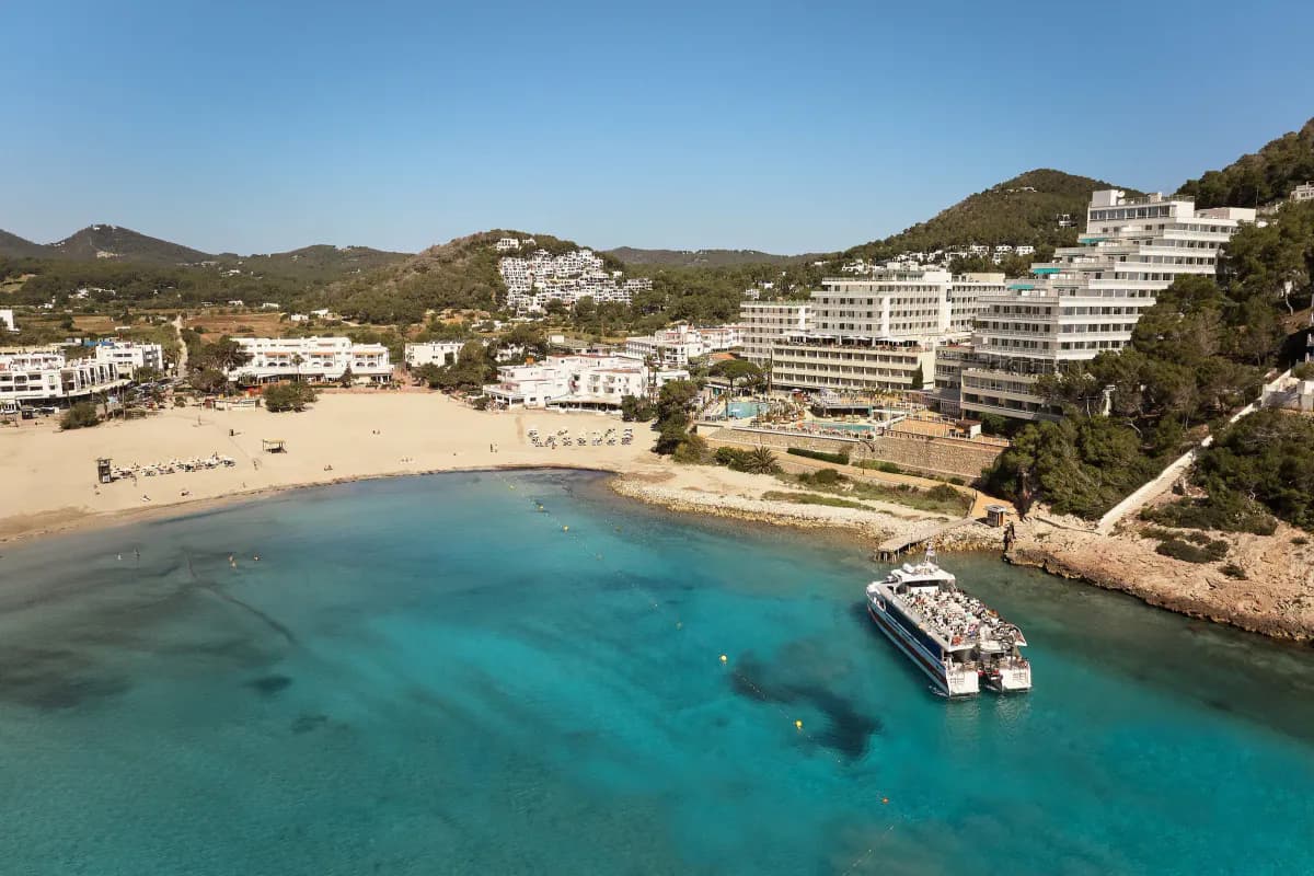 Zatoka Cala Llonga na Ibizie z lotu ptaka: piaszczysta plaża, turkusowa woda, statek wycieczkowy przy pomoście i białe hotele na zalesionych wzgórzach